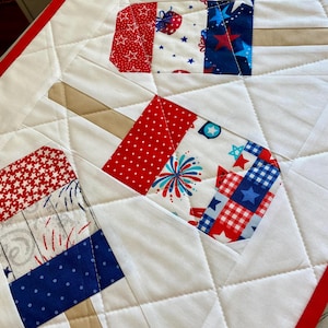May include: A quilted table runner featuring a popsicle design. The popsicles are made of red, white, and blue patterned fabric, with a white background and red border. The design includes stars, fireworks, and other patriotic motifs.