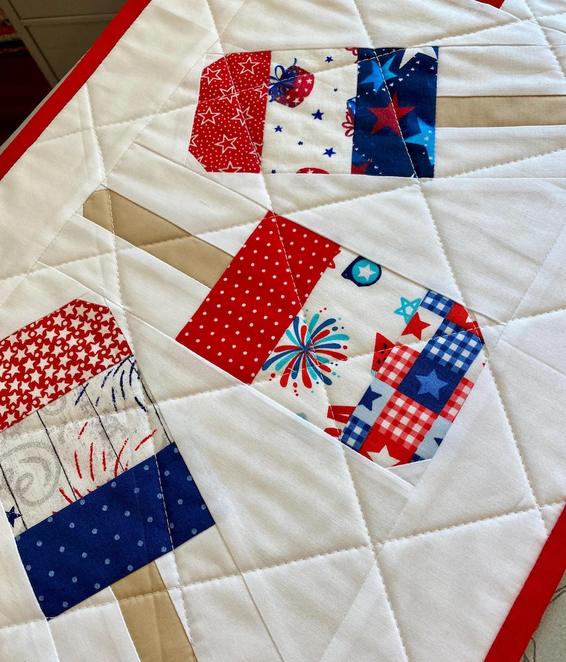 May include: A quilted table runner featuring a popsicle design. The popsicles are made of red, white, and blue patterned fabric, with a white background and red border. The design includes stars, fireworks, and other patriotic motifs.