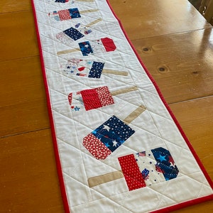 May include: A patriotic table runner featuring a repeating pattern of popsicle-shaped designs. The popsicles are made of red, white, and blue fabric with star and firework patterns. The runner is approximately 60 inches long and has a red border.