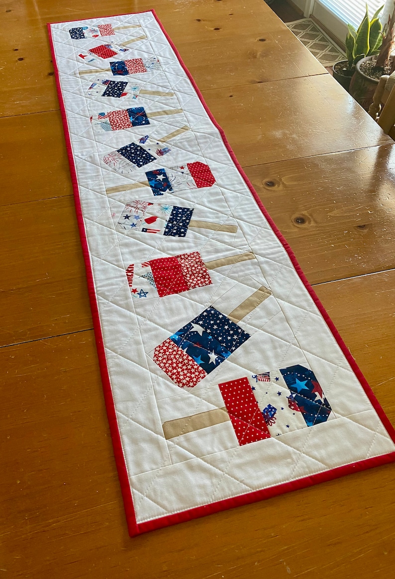 May include: A patriotic table runner featuring a repeating pattern of popsicle-shaped designs. The popsicles are made of red, white, and blue fabric with star and firework patterns. The runner is approximately 60 inches long and has a red border.