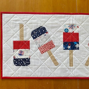 May include: A patriotic table runner featuring a repeating pattern of red, white, and blue popsicle designs. The popsicles are made of various patterned fabrics, including stars, stripes, and fireworks. The runner has a white background with a red border.