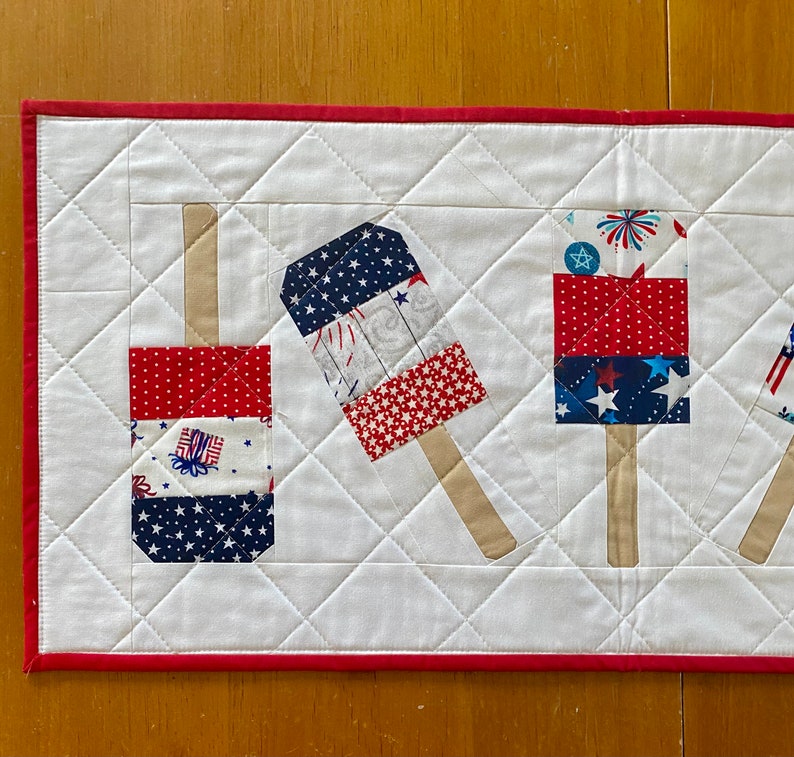 May include: A patriotic table runner featuring a repeating pattern of red, white, and blue popsicle designs. The popsicles are made of various patterned fabrics, including stars, stripes, and fireworks. The runner has a white background with a red border.