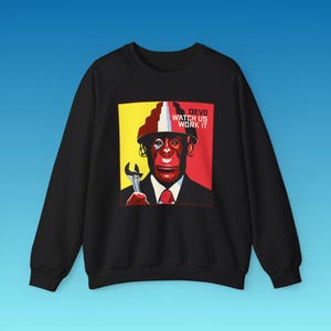 May include: A black long-sleeve sweatshirt featuring a graphic print of a primate wearing a red, white, and blue tiered hat and a suit with a red tie. The primate holds a silver wrench. The text reads DEVO WATCH US WORK IT in white.