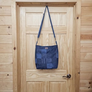 May include: A square-shaped crossbody bag constructed from a patchwork of denim fabric squares in varying shades of blue. The bag features an adjustable shoulder strap attached with metal hardware and a small rectangular brown leather-like brand tag.