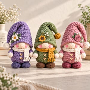 May include: Three handmade crochet gnomes in purple, green, and pink. Each gnome wears a tall pointed hat with a floral accent, a fringed scarf, and two small heart-shaped wooden buttons on the torso. They have round noses and white pom-poms.