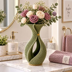 Op de afbeelding: Een saliegroene, geribbelde, zandlopervormige vaas met een boeket gehaakte bloemen in crème, mauve en paarse tinten, geaccentueerd met groene eucalyptusbladeren en kleine witte vulbloemen, staand op een aanrechtblad van marmer.