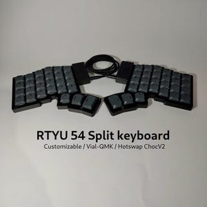 May include: A black split keyboard with grey keycaps, featuring the text RTYU 54 Split keyboard Customizable / Vial-QMK / Hotswap ChocV2. The device consists of two separate halves connected by a black cable, each with a grid of keys.