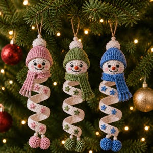 Könnte beinhalten: Drei gehäkelte Schneemann-Ornamente, die vertikal hängen. Jeder Schneemann hat einen weißen Kopf mit Karottennase, eine Strickmütze und einen passenden Schal. Darunter befindet sich ein langer, weißer Spiralkörper mit kleinen Sternapplikationen.