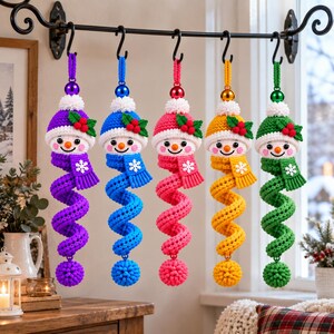 Könnte beinhalten: Fünf gehäkelte Schneemann-Ornamente hängen an einer schwarzen Metallstange. Jeder Schneemann hat eine farbenfrohe Mütze mit Stechpalmen, einen passenden Schal mit einer weißen Schneeflocke und einen langen, spiralförmigen Körper mit einem strukturierten Bommel.