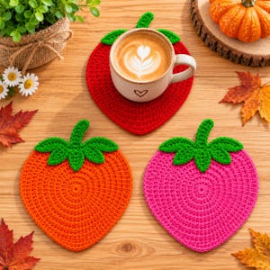 Könnte beinhalten: Drei erdbeerförmige Häkeluntersetzer in Rot, Orange und Pink, jeweils mit einem grünen Stiel und Blattaufsatz. Der rote Untersetzer trägt eine mit Kaffee gefüllte Keramiktasse, während die orangefarbenen und pinken Untersetzer auf Holz liegen.