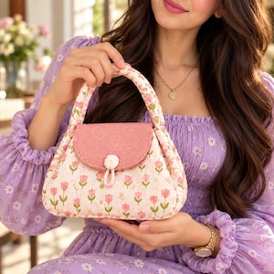 Könnte beinhalten: Eine gesteppte Stoffhandtasche mit cremefarbener Basis und einem wiederkehrenden Muster aus kleinen rosa Tulpen und grünen Stielen. Die Tasche hat eine einfarbige, strukturierte rosa Klappe, die mit einem stoffbezogenen Knopf verschlossen wird.