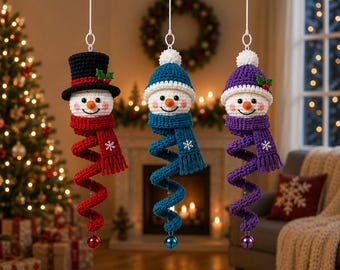 May include: Three crocheted snowman ornaments hanging vertically. Each features a white head with a carrot nose, rosy cheeks, and a colorful scarf that spirals downward into a tail ending with a small jingle bell. They wear hats in black, blue, and purple.