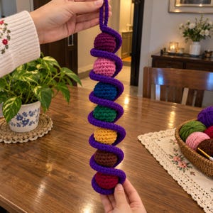 Könnte beinhalten: Ein vertikales Häkelspielzeug mit einer langen, violetten Spiralschnur, die sich um sieben bunte, kugelförmige Bälle windet. Die Bälle sind in einer Abfolge von Violett, Pink, Blau, Grün, Gelb, Braun und Rot angeordnet und aus Garn gefertigt.