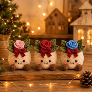May include: Three crocheted planters with smiling faces, pink blush, and small brown feet. Each features a central rose in pink, red, or blue, surrounded by green leaves and a small red bow. The planters are cream-colored with a textured yarn finish.