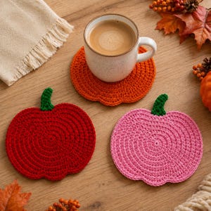 Könnte beinhalten: Eine Keramiktasse mit Kaffee steht auf einem gehäkelten Untersetzer in Form eines orangefarbenen Kürbisses. Zwei weitere kürbisförmige Untersetzer, einer in Rot und einer in Pink, jeweils mit einem grünen Stiel, ergänzen das Set.