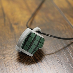 May include: A pendant shaped like a segmented hand grenade featuring a dark green grid pattern on a light gray base. The pendant is attached to a thin black cord that loops through a small gray ring at the top of the grenade-shaped charm.