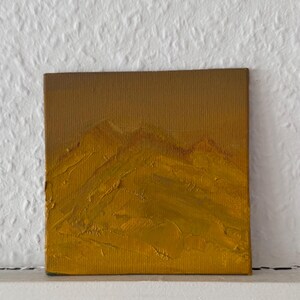 May include: A square canvas painting featuring a monochromatic landscape in shades of ochre and mustard yellow. The artwork displays a textured mountain range with thick, visible brushstrokes and a gradient sky that transitions from dark to light.