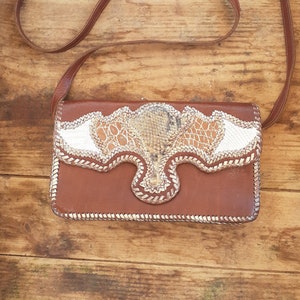 May include: A brown leather purse with a snakeskin pattern on the front flap. The purse has a long brown strap.