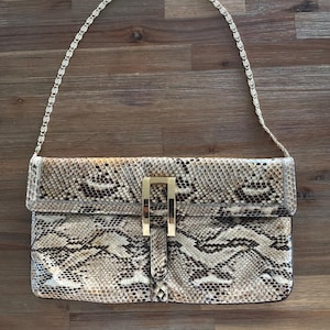 May include: A rectangular handbag with a snakeskin pattern in shades of brown, tan, and black. The bag features a gold-colored rectangular clasp and a chain strap. The bag is approximately 12 inches wide.