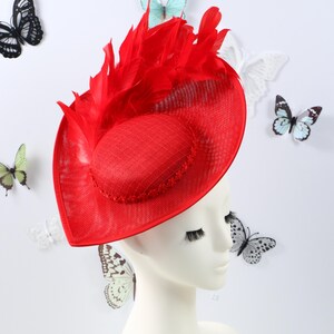 Puede incluir: Un tocado de color rojo brillante con una base estructurada de ala ancha hecha de material tejido. El sombrero está adornado con un grupo de plumas rojas erguidas y una sección central elevada de forma ovalada con un borde decorativo.