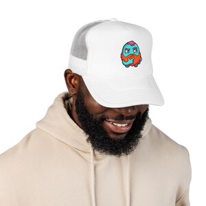 May include: A white trucker hat featuring a mesh back and a graphic print on the front. The graphic depicts a small, round, light blue creature with a bright orange beard, pink hair, and a thoughtful expression, resting its chin on its hand.