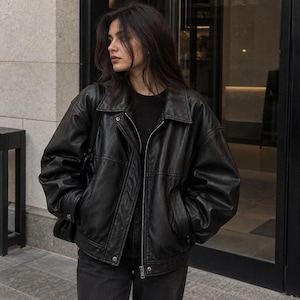 May include: An oversized black leather jacket featuring a classic collar, a full-length front zipper closure, long sleeves with buttoned cuffs, and two side pockets. The jacket is worn over a plain black shirt and dark-colored denim jeans.