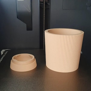 Peut inclure: Un récipient cylindrique de couleur pêche clair avec une texture nervurée verticale et un composant de base ou couvercle circulaire plus petit et bas, tous deux présentant un design cannelé cohérent, posés sur une surface sombre et plane.
