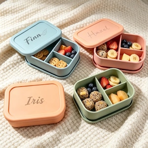 May include: Three rectangular silicone bento boxes with lids. One light blue box is labeled Finn, one peach box is labeled Hazel, and one orange box is labeled Iris. Each box features internal dividers and contains various snacks like fruit and crackers.