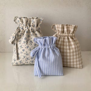 May include: Three fabric drawstring pouches featuring different patterns: one with a blue floral print on a cream background, one with blue and white vertical stripes, and one with a beige and white gingham check pattern, all with gathered tops.