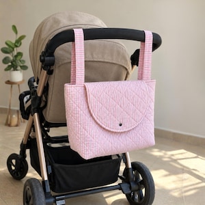 May include: A pink and white striped quilted stroller bag with a fold-over flap and a single snap closure. The bag features two long vertical straps that hang securely from the black handlebar of a beige and black baby stroller.