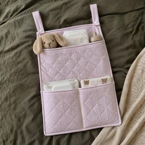 May include: A light pink quilted fabric hanging organizer with two tiers of pockets. The top tier holds a tan plush rabbit, a diaper, and a bottle. The bottom tier contains a pack of wipes and a small cloth with a bear print. It has two straps.