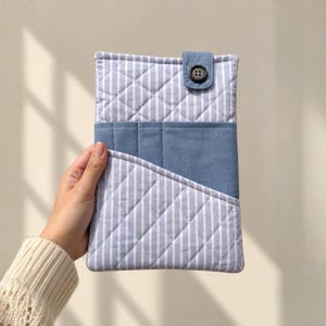 May include: A rectangular quilted fabric pouch featuring a light blue and white vertical striped pattern. The pouch includes a solid blue horizontal pocket section and a small blue fabric tab secured with a single black button at the top centre.