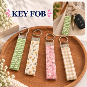 May include: Four fabric key fobs with silver-tone metal hardware and circular rings. The designs include a green oval pattern, a cream floral print, a pink daisy print, and a tan gingham check. One key fob is shown attached to a car key.