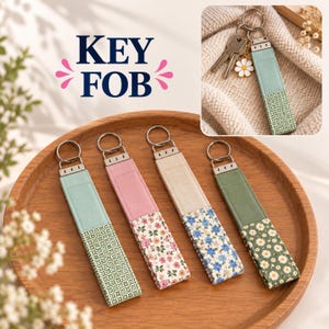 Op de afbeelding: Vier stoffen sleutelhangers met zilverkleurige metalen hardware en ringen. Elke hanger heeft een effen gekleurd bovendeel en een onderkant met bloemen- of geometrische patronen. Eén sleutelhanger is bevestigd aan metalen sleutels met een madeliefje-bedel.