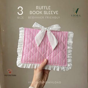 Puede incluir: Una funda para libro acolchada con rayas rosas y blancas, con un borde de volantes blanco y un lazo de tela blanco atado en el centro. El texto en la imagen dice 3 SIZE, RUFFLE BOOK SLEEVE, BEGINNER FRIENDLY, VIORA PATTERN y PDF PATTERN.