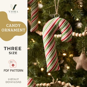 Può includere: Un ornamento a forma di bastoncino di zucchero in tessuto con un motivo a righe diagonali rosse, bianche e verdi. L'ornamento è appeso a un filo di iuta fissato a un anello di metallo argentato. Il testo Viora Pattern Candy Ornament è visibile.