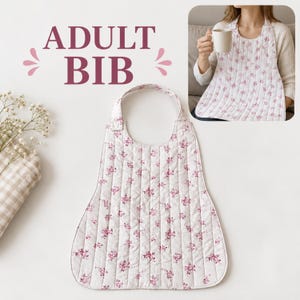 Può includere: Una bavaglia per adulti in tessuto trapuntato bianco con un piccolo motivo floreale rosa. La bavaglia presenta uno scollo rotondo e una chiusura a pressione sulla spalla. Il testo ADULT BIB è stampato in grandi lettere marrone scuro.