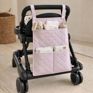 Könnte beinhalten: Ein hellrosa gesteppter Kinderwagen-Organizer mit dezentem Karomuster, ausgestattet mit zwei oberen Riemen mit Druckknöpfen, einer großen oberen Tasche und zwei kleineren unteren Taschen, die ein Stofftier, Windeln und eine Flasche enthalten.