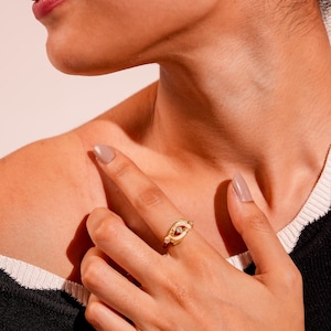 Puede incluir: Un anillo de tono dorado que presenta un diseño central de forma ovalada con una pequeña piedra clara y brillante engastada en el medio. La banda tiene una superficie texturizada y ligeramente irregular, y se lleva puesta en un dedo.