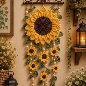 Op de afbeelding: Een wandhanger met een grote centrale gehaakte zonnebloem met een donkerbruin hart en gele bloemblaadjes, opgehangen aan een donkere metalen stang. Kleinere gehaakte zonnebloemen en groene blaadjes zitten aan hangende koorden met houten kralen.