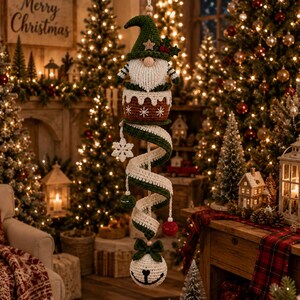 May include: A hanging crochet Christmas decoration featuring a gnome in a green hat sitting in a brown pudding-shaped base. A long, spiraling green and white ribbon extends downward, accented with a white snowflake, small spheres, and a white ball ornament.