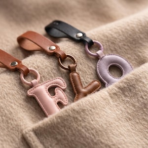 Könnte beinhalten: Drei Schlüsselanhänger aus Leder mit den Buchstaben F, Y und O. Jeder Anhänger hat ein Lederband mit einer silberfarbenen Metallniete, das an einem Ring befestigt ist, an dem ein gepolsterter Lederbuchstabe in Hellrosa, Braun oder Flieder hängt.