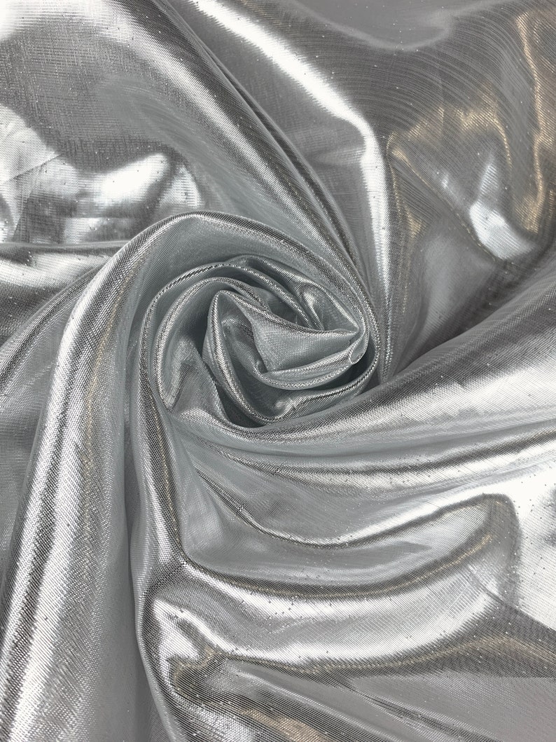 May include: Silver metallic fabric with a shimmering, iridescent finish. The fabric is draped and folded, showcasing its texture and drape.