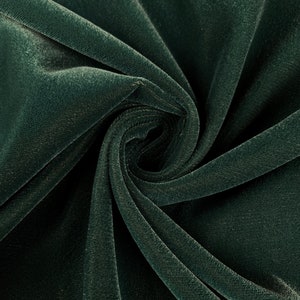 May include: Close-up of a dark green velvet fabric with a soft, plush texture. The fabric is folded and draped, showcasing its smooth surface and subtle sheen.