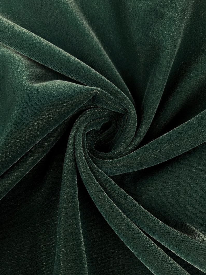 May include: Close-up of a dark green velvet fabric with a soft, plush texture. The fabric is folded and draped, showcasing its smooth surface and subtle sheen.
