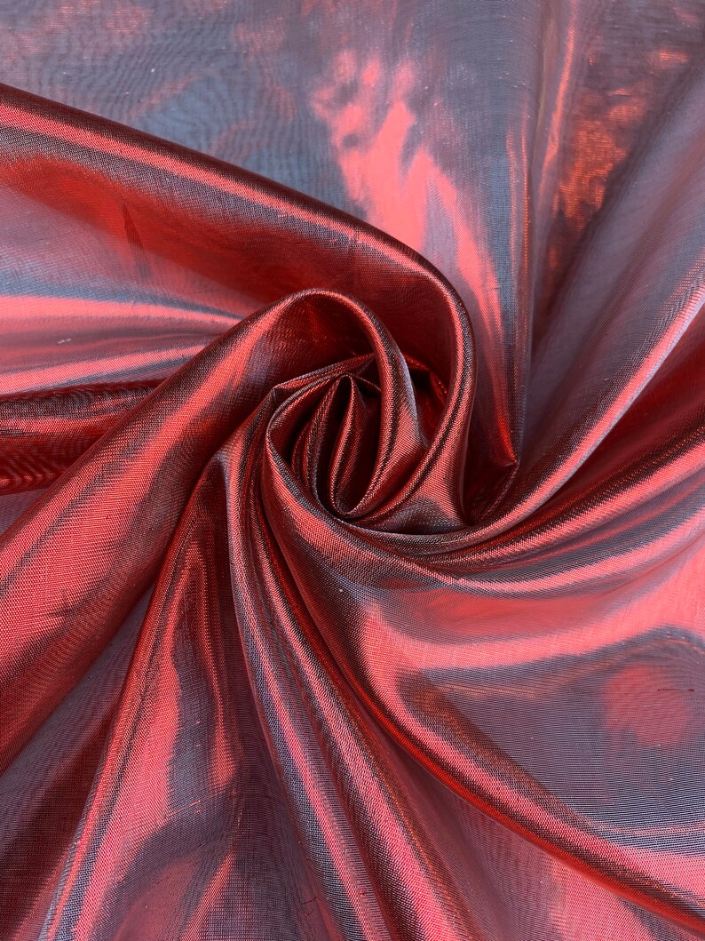 May include: A close-up of a red iridescent fabric with a silky sheen. The fabric is draped and folded, creating a soft, flowing texture.
