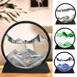 May include: A circular sand art display featuring a black frame and base. The glass contains layered sand forming mountain landscapes in black, white, and gold. Additional versions are shown in green and white, and blue and white color schemes.