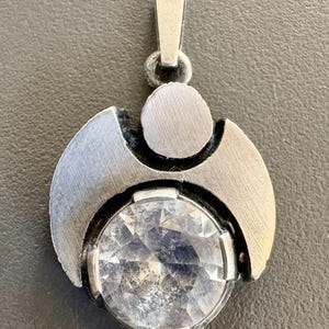 May include: A silver-toned metal pendant featuring a brushed finish, a circular top element, and a large, round, faceted clear stone set at the base. The pendant includes a vertical bail at the top for attachment to a chain or cord.