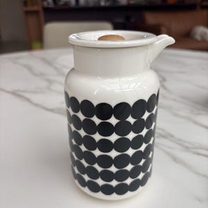 Op de afbeelding: Een witte keramische kan met een herhalend patroon van zwarte cirkels. De kan heeft een cilindrisch lichaam, een korte tuit aan de zijkant en een bijpassend wit deksel met een kleine, ronde houten knop als handvat.