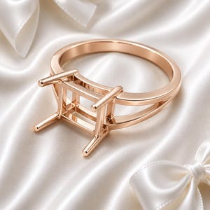 May include: A rose gold ring setting featuring a square-shaped head with four prongs designed to hold a gemstone. The band has a split shank design that connects to the central mounting, showcasing a polished metallic finish throughout the piece.